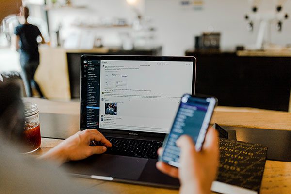 Persona usa portátil mientras sostiene un teléfono móvil en cafetería, con pantalla de chat abierta y bebida sobre la mesa.