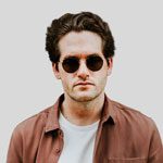 Retrato de un hombre moreno con gafas de sol redondas, chaqueta marrón, camisa blanca y fondo gris.