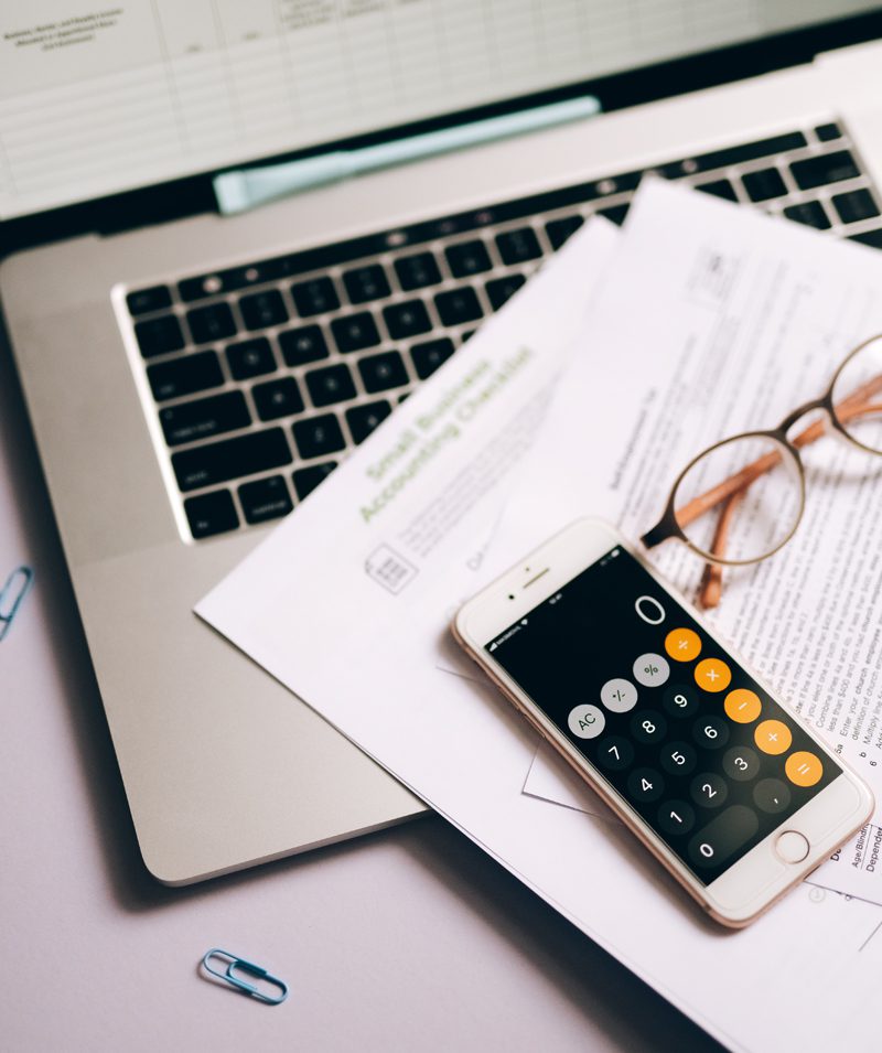Teléfono con calculadora sobre documentos financieros y portátil, junto a gafas y clip metálico en escritorio de trabajo.