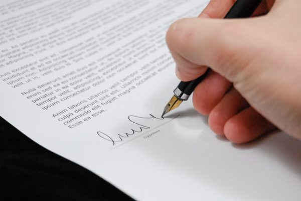 Mano sostiene pluma estilográfica mientras firma documento impreso sobre mesa oscura, con texto del contrato visible alrededor.