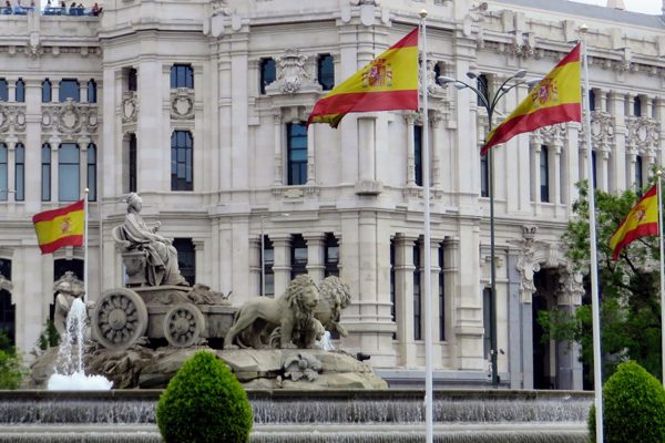 Fuente con escultura de carro tirado por leones frente a edificio histórico, con banderas de España ondeando en plaza urbana.