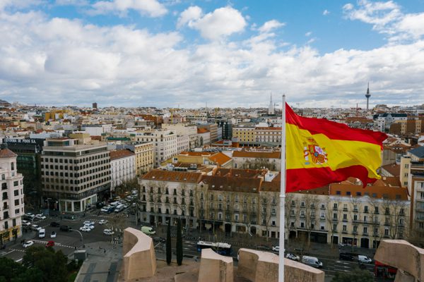 Bandera de España ondea sobre una ciudad con edificios históricos y cielo azul con nubes, vista panorámica urbana.